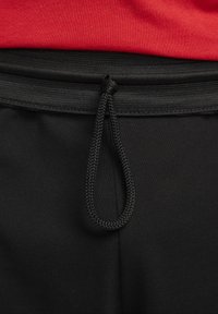 Pantalon noir à cordon avec une boucle nouée à la taille, associé à une chemise rouge rentrée.