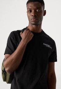 Black cotton t-shirt with crew neckline, featuring white embroidered text "Espresso Awakening," worn with an olive green backpack.