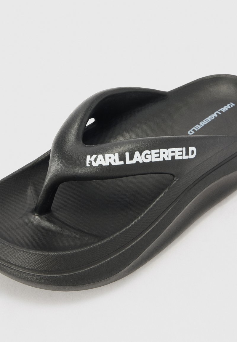 Czarna klapek z fakturowanym wykończeniem, z wyraźnym podniesionym paskiem na palce i białym logo "KARL LAGERFELD" po boku.