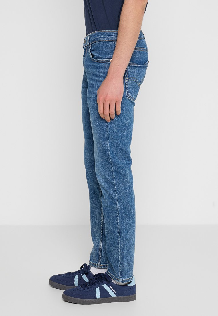 Jeans de denim azul con un corte recto, que presentan un diseño de cinco bolsillos y una sutil textura desgastada. Llevados con zapatillas de deporte azul marino con detalles en azul claro.