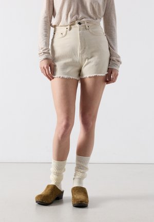 Person iført beige højtaljede denimshorts med frynser, cremefarvede løsthængende sokker, brune ruskindsclogs og en lys, langærmet top, stående indendørs.
