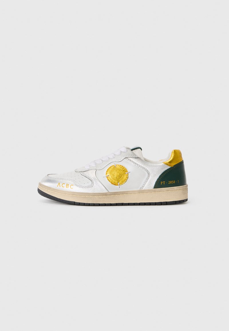 Zapatilla de piel blanca con acentos metálicos plateados, talón verde, cuello amarillo, logo circular y texto impreso en el lateral y la lengua.
