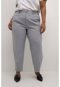 Pantaloni grigi chiari su misura con vestibilità dritta, dotati di chiusura a bottone, pieghe leggere e spacco laterale all'orlo, abbinati a scarpe nere.