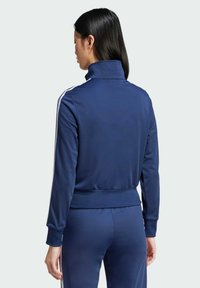 Marineblaues Sportjackett aus glatter Stoffqualität, mit hohem Kragen, Reißverschluss und weißen Streifen entlang der Ärmel.
