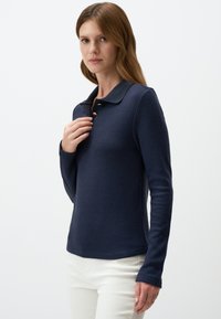 Marineblauwe langemouwen polo-shirt met een kraag en een knoopsluiting met drie knopen, gemaakt van een textuurstof en een getailleerd ontwerp, gecombineerd met witte broek.