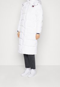 Vit pufferjacka med huva, med quiltade sektioner, framzipper och en liten logotypapplikation. Kombinerad med mörka jeans och vita sneakers.