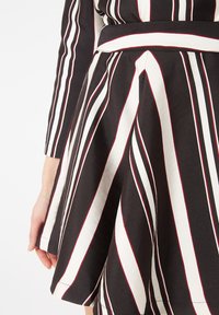 Primer plano de un vestido con rayas verticales negras, blancas y rojas, cintura ajustada y falda acampanada, mostrando un brazo y mano parcialmente visibles.