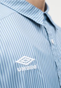 Μπλε-άσπρο ριγέ πουκάμισο με γιακά και το άσπρο λογότυπο Umbro κεντημένο στο στήθος, που φοριέται από ένα άτομο.