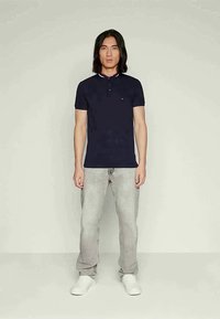 Man som står klädd i en marinblå kortärmad polo, ljusgrå jeans och vita sneakers mot en enkel ljus bakgrund.