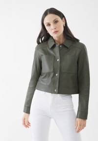 Giacca in pelle verde oliva tagliata con colletto a punta, due tasche frontali e bottoni a pressione argentati. Indossata con jeans bianchi.
