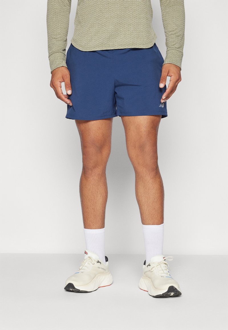 Homme portant un short de sport bleu marine, des chaussettes blanches montantes et des chaussures de course blanches, debout devant un fond clair uni.