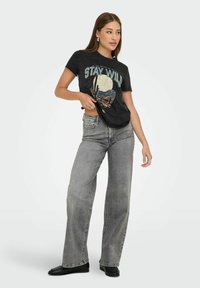 Zwarte grafische T-shirt met een woestijnscene en de tekst "STAY WILD," gecombineerd met lichtgrijze wijde spijkerbroek en zwarte platte schoenen.