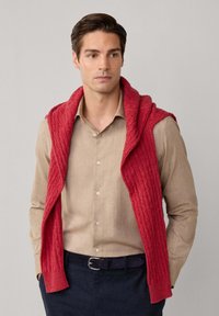 Rode gebreide mouwloze cardigan met geribbelde structuur, over een beige blouse met lange mouwen gedrapeerd, gecombineerd met donkerblauwe broek en een zwarte riem.