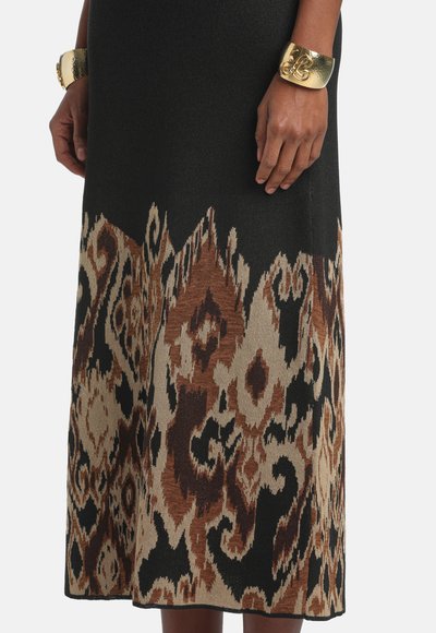 Čierne šaty s hnedými a béžovými abstraktnými vzormi pri okraji, nosené so širokými zlatými manžetovými náramkami na oboch zápästiach.
