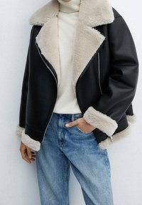 Veste en cuir noir avec un col et des poignets en fausse fourrure beige, fermeture éclair, portée par-dessus un col roulé crème et un jean bleu.