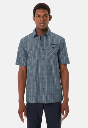 Uomo in piedi che indossa una camicia a quadri blu a maniche corte con taschino sul petto e pantaloni scuri davanti a uno sfondo semplice.