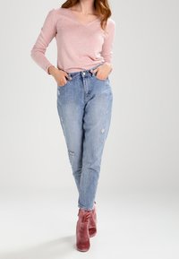 Jasnoróżowy sweter z dekoltem w serek, podarte jasnoniebieskie dżinsy z naszywkami oraz różowe botki za kostkę. Strój charakteryzuje się casualowym krojem i miękkimi teksturami.