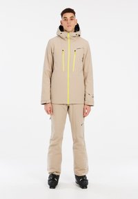 Beige Skijacke mit schwarzem Futter, Reißverschluss vorne und zwei Brusttaschen, die in Gelb hervorgehoben sind. Kombiniert mit beigen Skihosen und schwarzen Skischuhen.