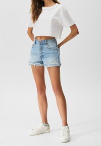 T-shirt bianca cropped abbinata a pantaloni corti in denim azzurro chiaro con orli sfrangiati e tasche. Sneakers bianche con dettagli verdi completano il look.