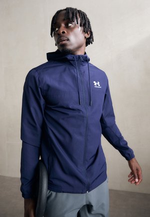 Tamsiai mėlynas lengvas bliuzonas su gaubtu, visas ilgis užtrauktuku, elastinėmis rankovėmis ir Under Armour logotipu kairėje krūtinės pusėje.