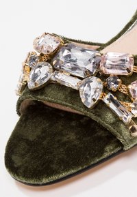Samettivihreä avokärkinen kenkä, jossa on suuria läpinäkyviä ja vaaleanpunaisia jalokivikoristeita remmissä.