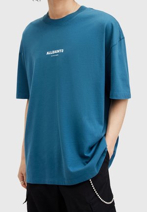 Personne portant un t-shirt ample à manches courtes bleu avec le texte blanc inversé « ALLSAINTS » et un pantalon noir avec une chaîne argentée sur un fond gris clair.