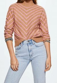 Suéter marrón con franjas de chevron rosas, cuello redondo y mangas tres cuartos. Lleva puesto sobre jeans azules claros de talle alto con un corte clásico.