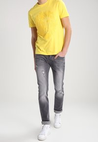 Žluté bavlněné tričko s grafikou lva, zkombinované s bledě šedými džíny se zdrsněnými detaily. Bílé tenisky dokončují outfit.