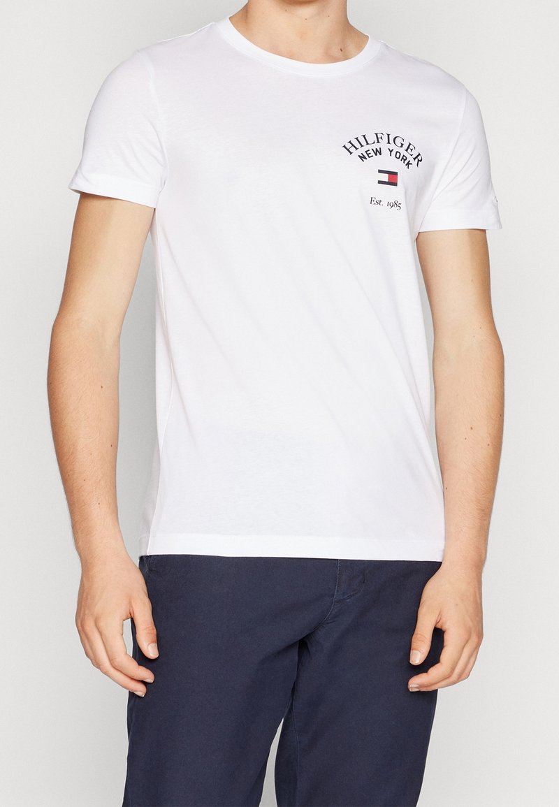 Camiseta blanca de manga corta hecha de algodón, con cuello redondo y un estampado de logo en el pecho en colores azul marino y rojo.