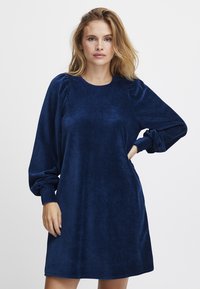 Navyblaue Cordkleid mit rundem Ausschnitt, langen Puffärmeln und lockerer Passform. Der Stoff hat eine weiche Textur mit vertikalen Rippungen.