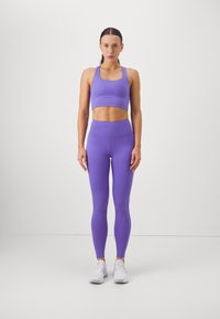 Μωβ αθλητική εμφάνιση που περιλαμβάνει ένα εφαρμοστό crop top και ψηλόμεσα κολάν, κατασκευασμένα από ελαστικό υλικό. Συνδυασμένο με λευκά αθλητικά παπούτσια.