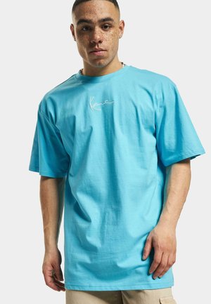 Camiseta de algodón turquesa con mangas cortas y corte relajado. Presenta un simple logo blanco en el pecho. Textura suave y sin patrones.