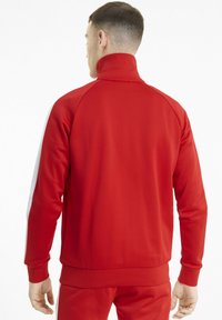 Veste de survêtement rouge avec col haut, poignets côtelés et ourlet. Présente des bandes latérales blanches et une texture lisse. Le design met l'accent sur une coupe athlétique.