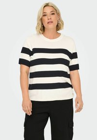 Top a maniche corte a righe in bianco e blu marino, con righe orizzontali, scollatura rotonda e vestibilità comoda. Sotto si indossano pantaloni neri.