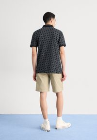Sort polo-shirt med et subtilt geometrisk mønster, parret med beige shorts og hvide høj-top sneakers. Ensfarvet baggrund.