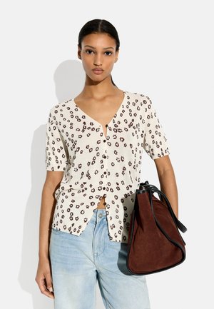 Kobieta ubrana w biały, krótki rękaw, zapinany na guziki top z brązowym kwiatowym wzorem, jasnoniebieskie dżinsy, trzyma dużą brązową zamszową torebkę z czarną lamówką.