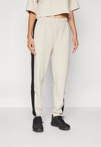 Beige joggeri ar melnām sānu svītrām, elastīgu jostasvietu un sašaurinātām kājām, kopā ar melnu sporta apavu pāri. Maiga, gluda auduma tekstūra.
