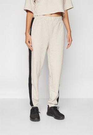 Beige joggers with black side stripes, elastic waistband, and tapered legs, paired with black athletic shoes. Soft, smooth fabric texture.
