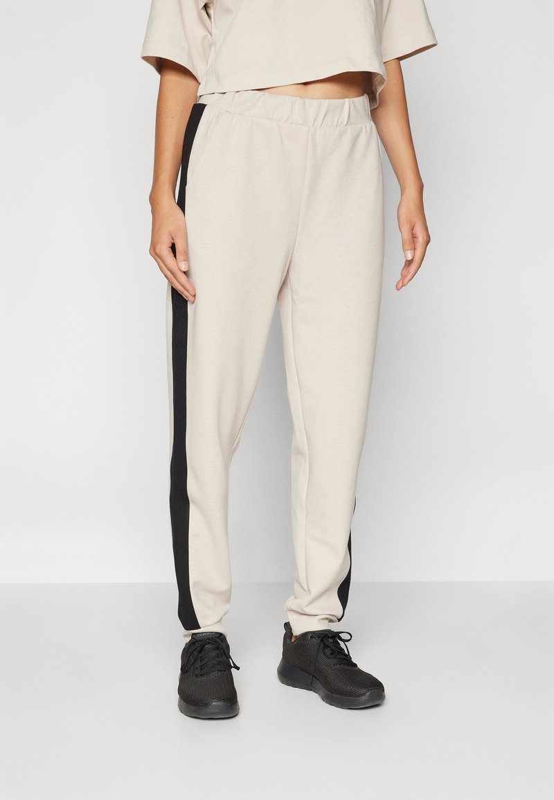 Beige joggeri ar melnām sānu svītrām, elastīgu jostasvietu un sašaurinātām kājām, kopā ar melnu sporta apavu pāri. Maiga, gluda auduma tekstūra.