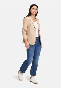 Beżowa marynarka z dwoma guzikami i dwiema przednimi kieszeniami, zestawiona z białym topem na ramiączkach i niebieskimi dżinsami, dopełniona białymi sneakersami.
