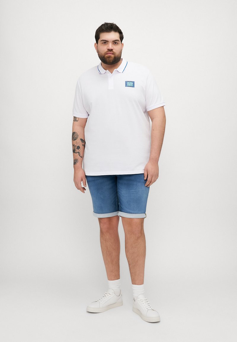 Camisa polo blanca con un logo azul, con un cuello clásico y un ribete azul. Combinada con pantalones cortos de mezclilla azul claros y zapatillas blancas.