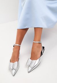 Scarpe slingback argento con punta affusolata, dettaglio a fiocco rigido prominente e cinturino alla caviglia. Finitura metallica liscia, design con tacco piatto.