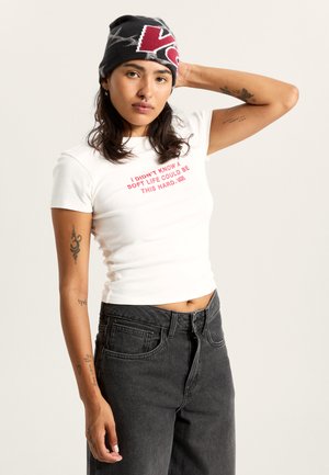 Wit katoenen t-shirt met rode tekst, gecombineerd met hoogwater zwarte denim jeans. Een gebreide zwarte muts met rood-wit logo.