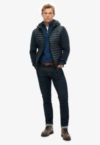 Herenoutfit met een zwarte puffer vest, marineblauw trui, donkere spijkerbroek, bruine riem en bruine laarzen; met gladde texturen en een casual ontwerp.
