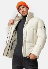 Puffjakke i blødt, off-white stof med høj krave, lynlåslynlås og logo-patch. Kombineret med en mørk turtleneck og orange hue.