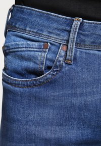 Närbild av en ficka på blå denimjeans med kopparnitar och ett bälteshål, bärs med en svart topp.