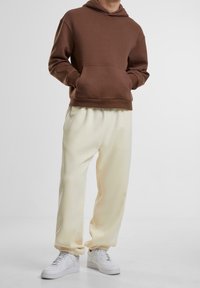 Persona con una sudadera marrón, pantalones de chándal color crema y zapatillas blancas, de pie con las manos en los bolsillos frente a un fondo liso.