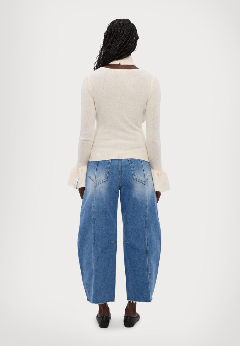 Pull en tricot beige avec un corsage ajusté, détails de manches évasées, associé à un jean en denim bleu clair à taille haute et jambes larges, présentant une finition délavée.