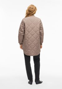 Brązowa pikowana długa kurtka z okrągłym dołem, ozdobiona zaokrąglonymi wzorami przeszyć i luźnym krojem, zestawiona z czarnymi spodniami o prostych nogawkach.