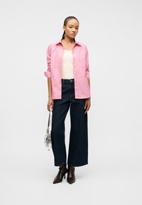 Γυναίκα με πλεξούδες, που φοράει ροζ ριγε πουκάμισο πάνω από λευκό τοπ, σκούρα τζιν με φαρδιά μπατζάκια, μαύρες μπότες, κρατώντας ασημί τσάντα.